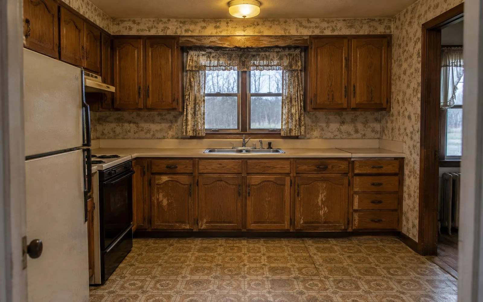 A dated kitchen with worn oak cabinets, floral wallpaper, laminate counters and patterned vinyl flooring
